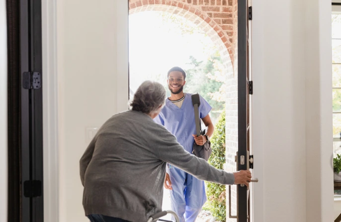 Male nurse in light blue scrubs greeting a patient at the entrance of a home